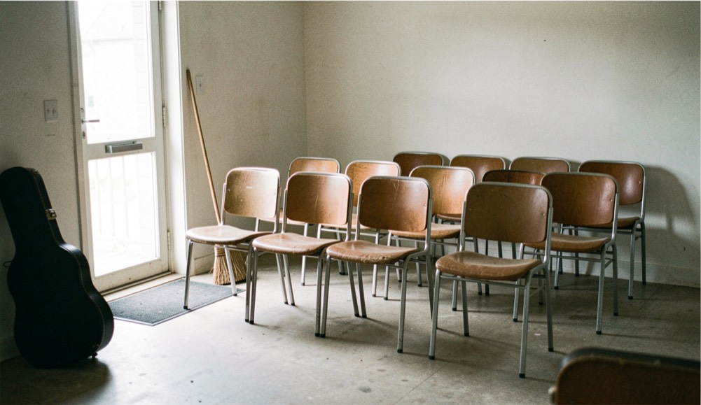 An empty small room in daylight with chairs in rows of six, a broom leaning by the door and a guitar case on the floor